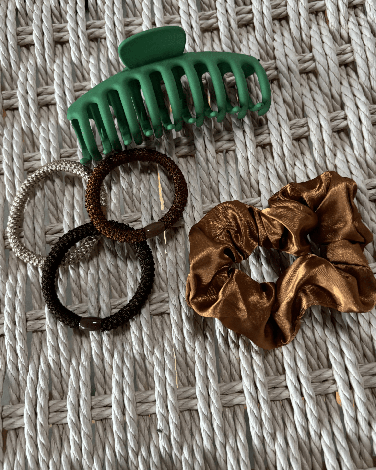 Grøn hårklemme, brun satin scrunchie og tre hårelastikker i jordfarver på flettet baggrund.