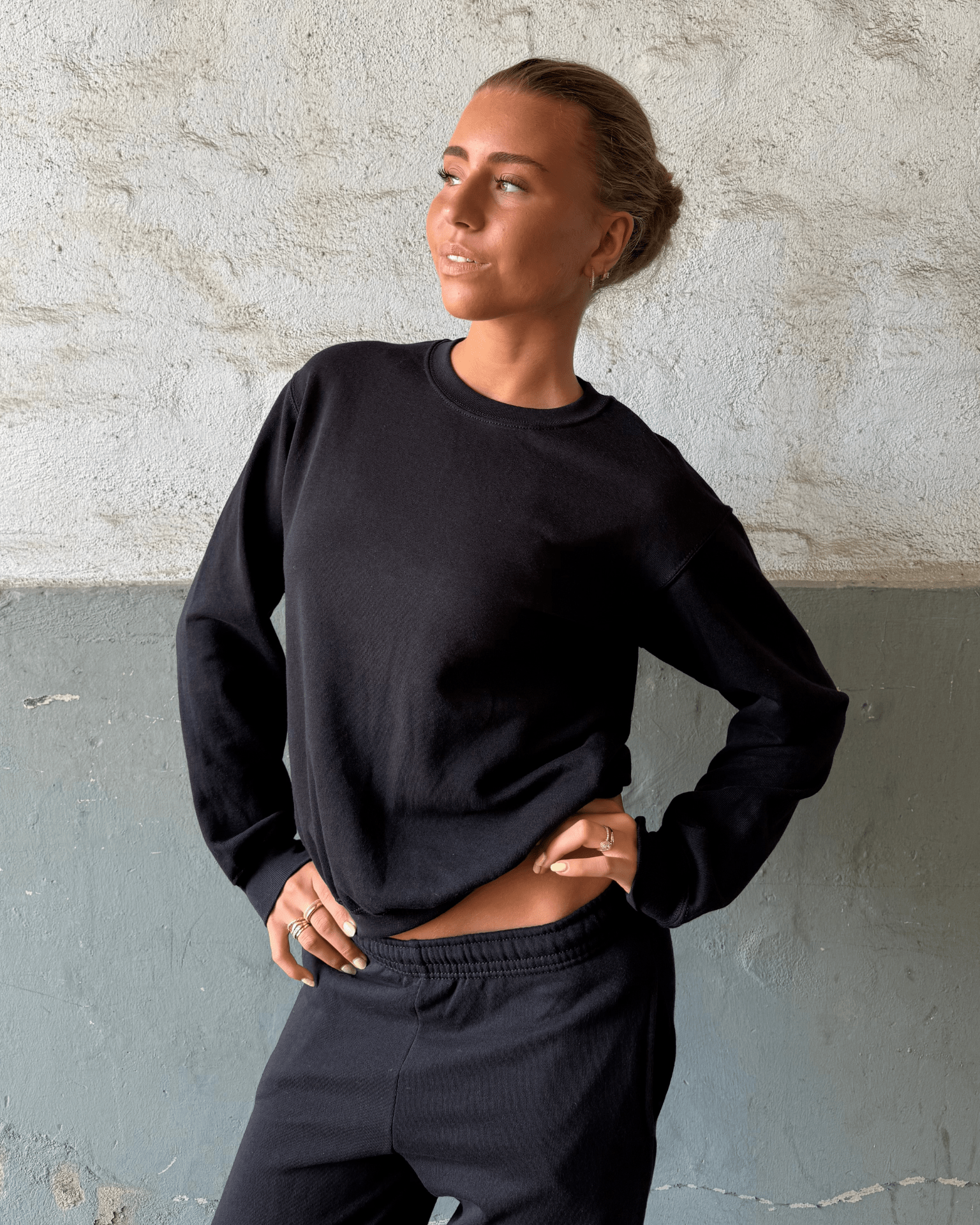 Kvinde i sort Fruit sweatshirt og matchende joggingbukser mod en rå væg