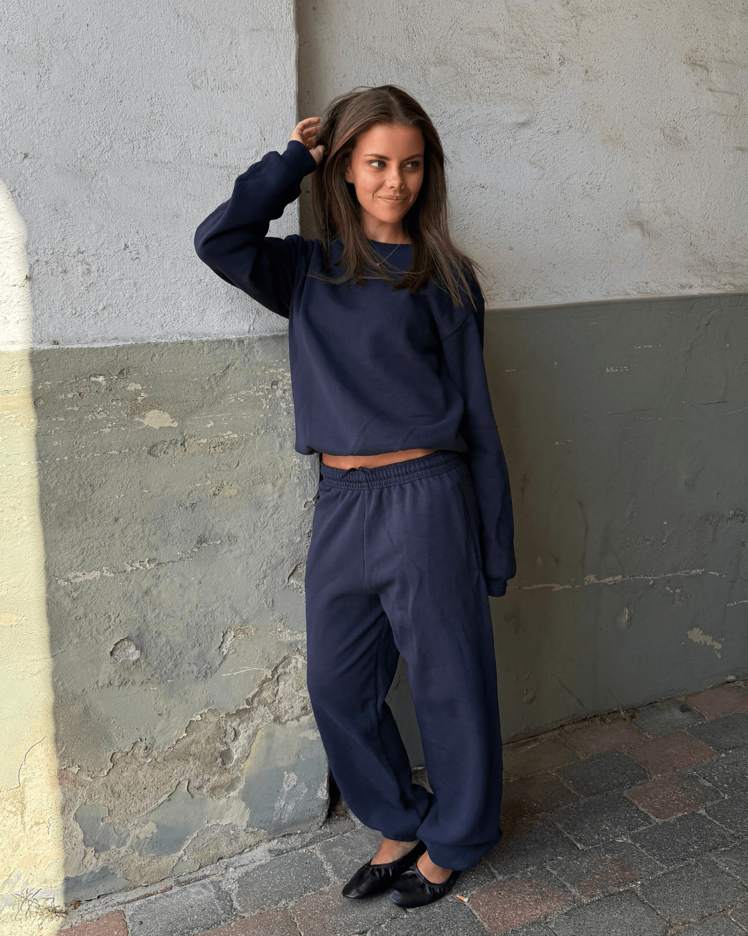 Kvinde i mørkeblå Fruit sweatshirt og matchende joggingbukser mod en rå væg