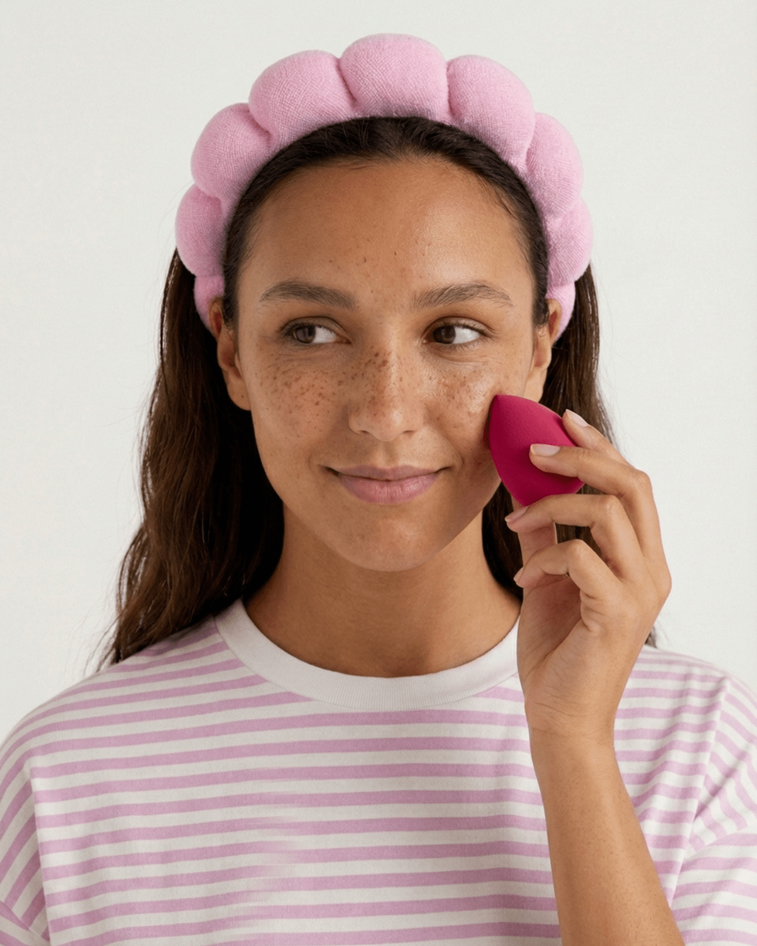 Kvinde med lyserødt skumpandebånd, der bruger en pink beautyblender mod kinden.