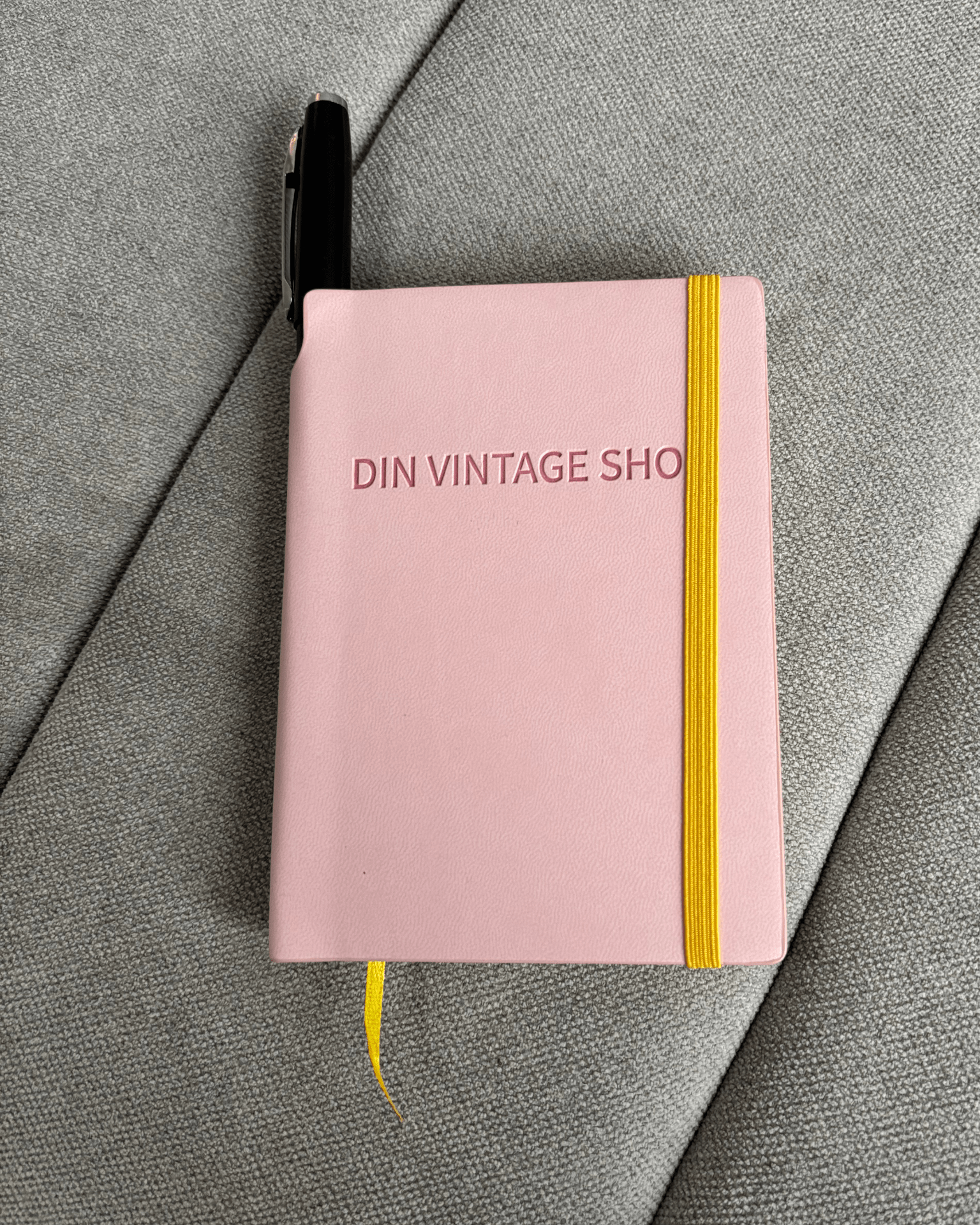 Lyserød Din Vintage Shop planner med gul elastik og sort pen på gråt stof