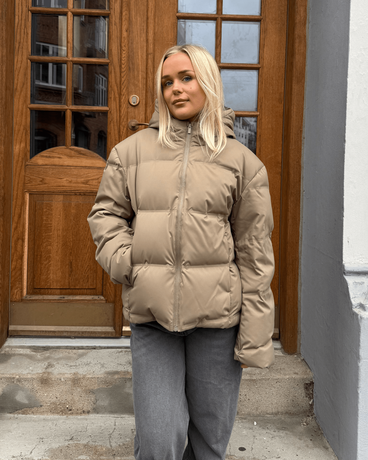 Taupe Isa dunjakke med hætte og lynlås på model foran en trædør