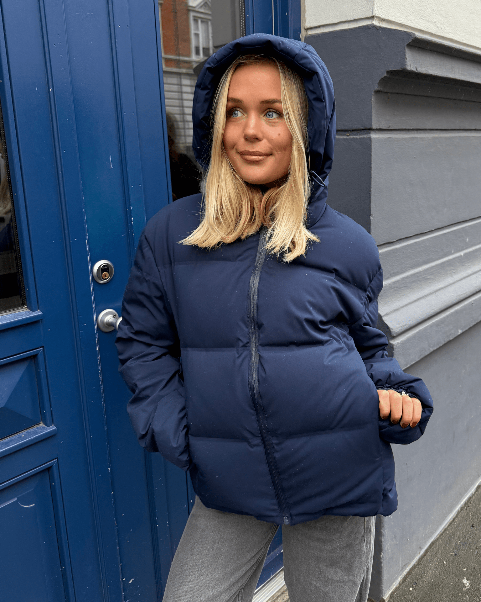 Model i navyblå Isa dunjakke med stor hætte foran en blå dør