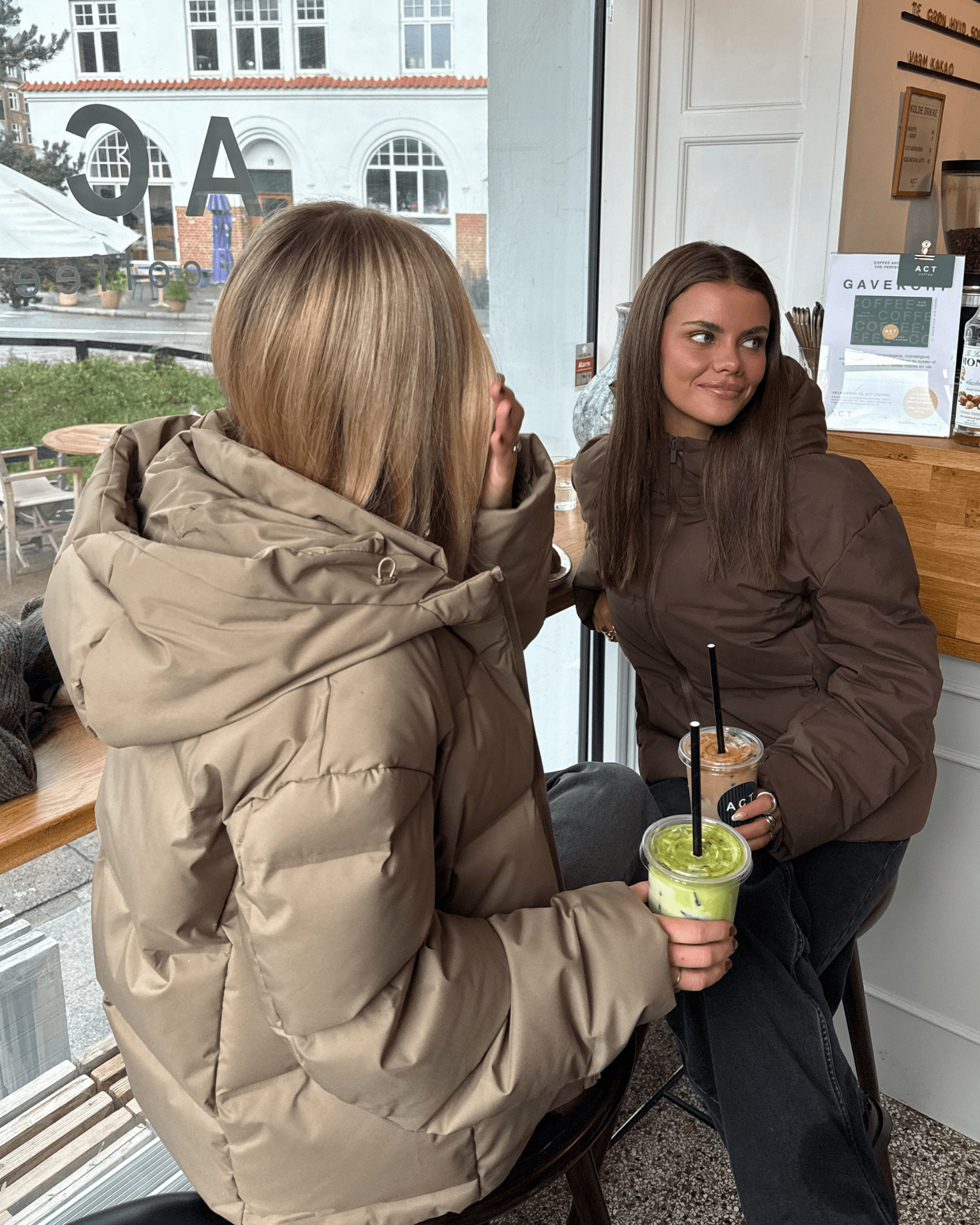 To kvinder i brune Isa dunjakker med hætte på café